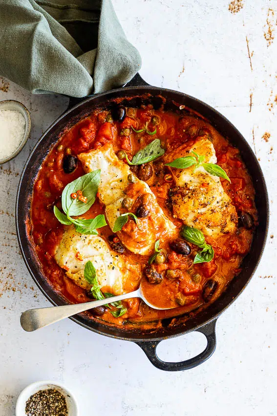 Seared fish in puttanesca sauce in pan with spoon on table top.