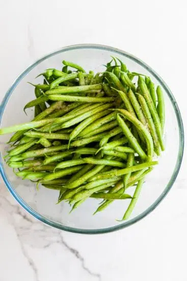 Trimmed green beans in bowl with olive oil and seasonings.