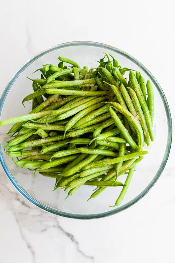 Trimmed green beans in bowl with olive oil and seasonings.