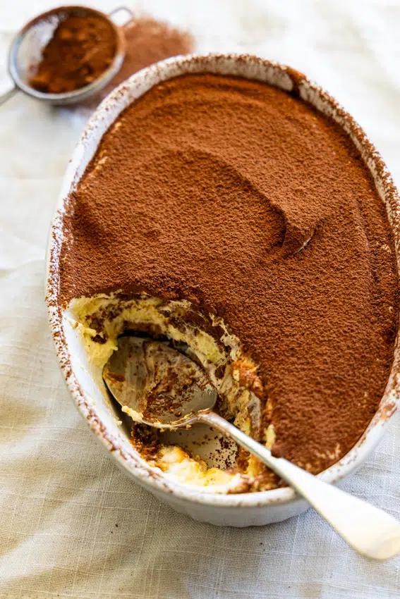 Layers of creamy tiramisu topped with cocoa powder in a serving dish with spoon.