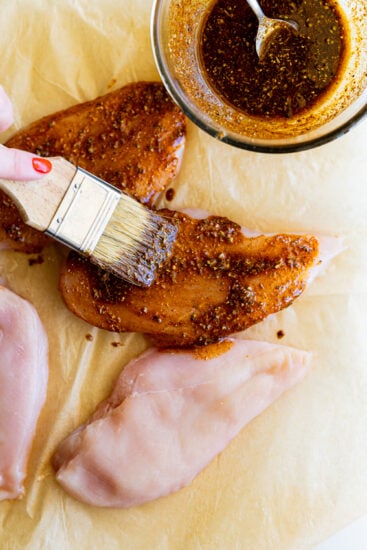 Seasoning and oil brushed over raw chicken breasts.