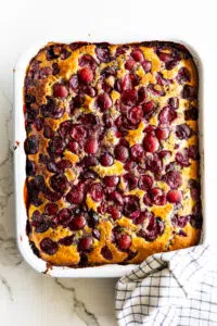 Baked cherry cobbler in a white baking dish with a checkered cloth on a marble surface.
