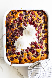 Baked cherry cobbler in white baking dish with vanilla ice cream and a checkered cloth.