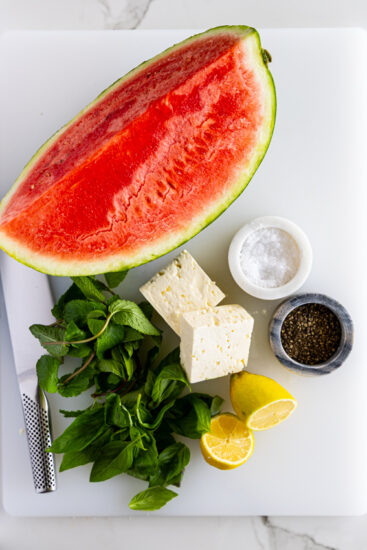 Ingredients for watermelon salad with feta cheese on a white surface. Fresh watermelon, feta cheese, fresh basil, lemon, salt and pepper with a chef's knife on white cutting board.