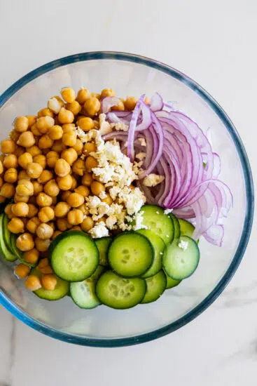 Cucumber, chickpeas, feta cheese and red onion in a glass bowl.