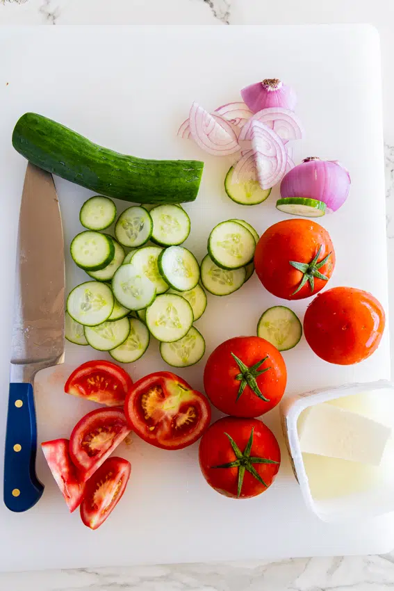 Ingredients for cucumber, tomato and feta salad on a white cutting board with a chef's knife.