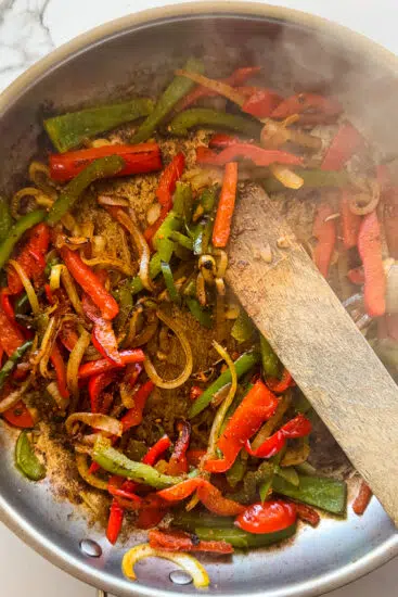 Sauteéd peppers and onions in a frying pan with a wooden spoon.