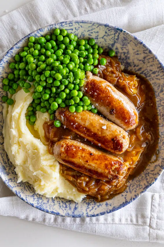 Bangers and mash with onion gravy and frozen peas served in a bowl.