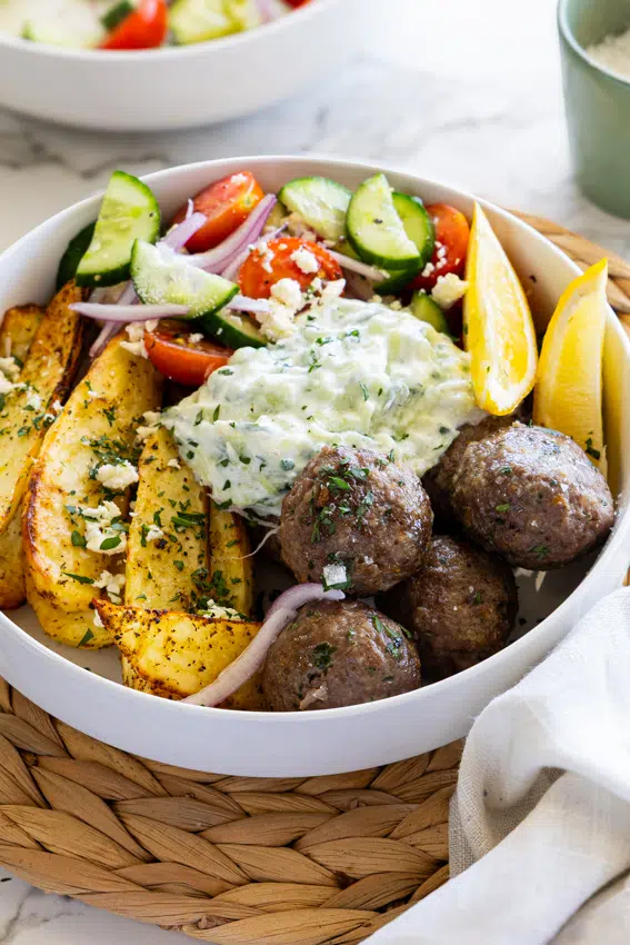 Juicy Greek meatballs with lemon roasted potatoes and Greek salad and tzatziki in a white bowl.